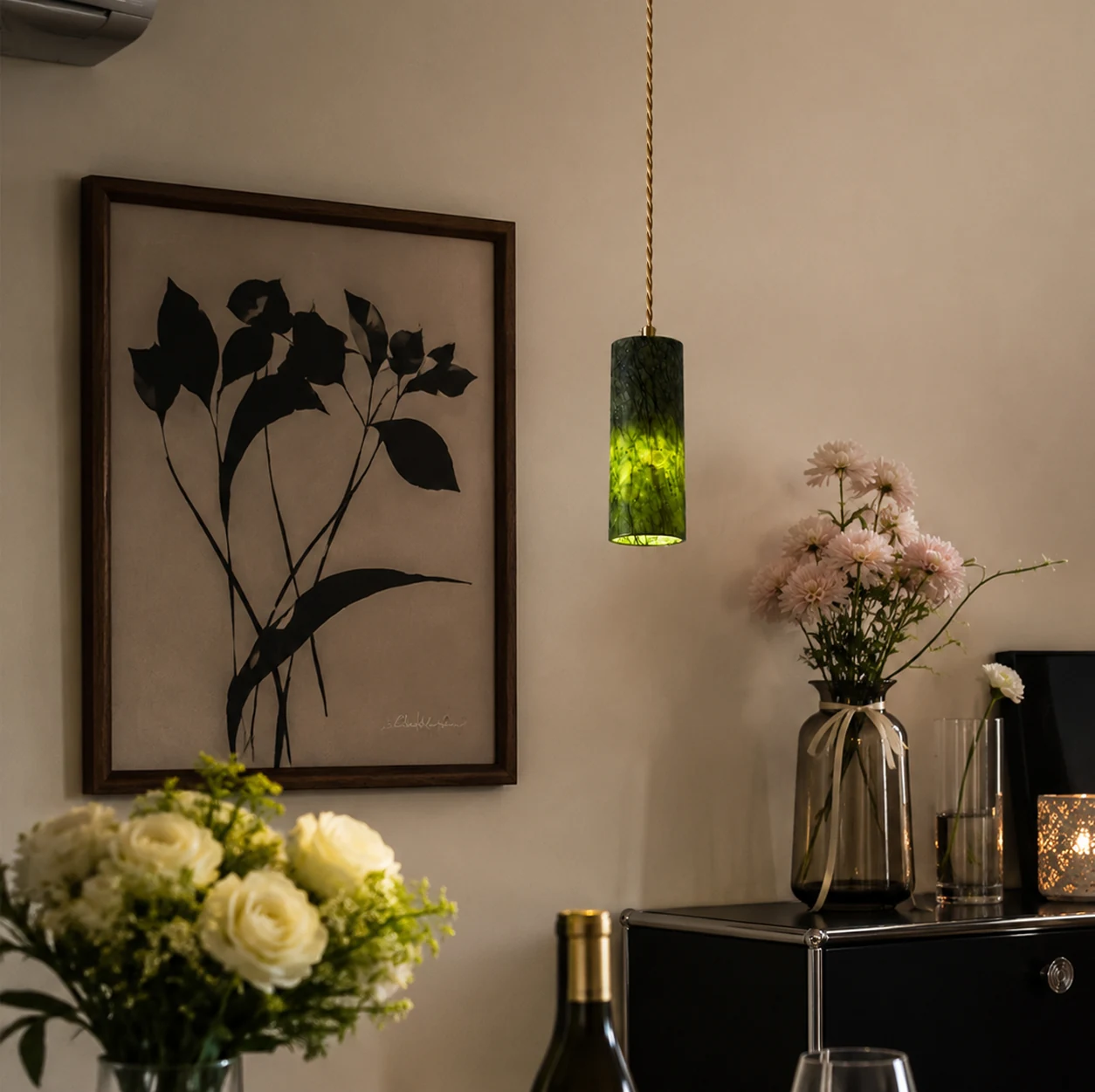 Single green jade cylinder pendant light glowing beside framed botanical wall art and flowers